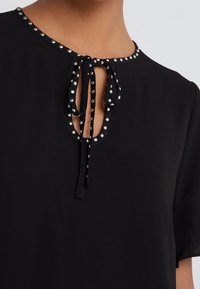 Sort bluse med rund halsudskæring, der har en nøglehul-detalje og en bindelukning. Accentueret med sølvnitter langs halsudskæringen.