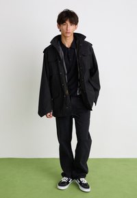 Jachetă neagră cu fermoar, dotată cu multiple buzunare frontale, purtată peste un tricou polo negru, asortată cu blugi negri și adidași negri. Fundal verde.