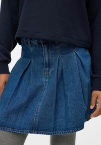 Falda de mezclilla con diseño plisado, color azul y costuras visibles. Llevada con un sudadera oscura y leggings grises.