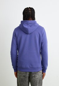 Haut à capuche violet avec une poche kangourou et un cordon de serrage. Tissu doux, poignets et ourlet côtelés. Porté avec un jeans texturé gris.