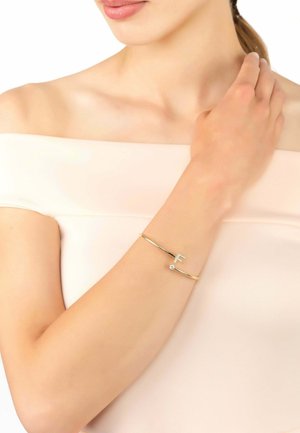 Bracciale a polsino in oro con un dettaglio a forma di "F" e una piccola pietra preziosa trasparente, indossato su un braccio nudo contro uno sfondo chiaro e neutro.