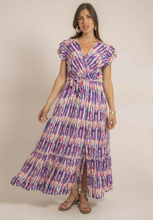 Femme portant une robe maxi colorée à motifs, manches courtes, ceinturée, avec une fente à l'avant, et des sandales à clous, debout devant un fond neutre.