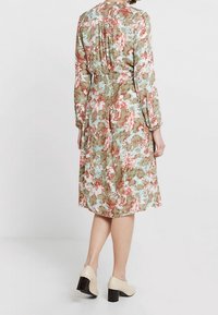 Robe imprimée florale en vert, rose et bleu avec des manches longues, taille cintrée et détail de pli dans le dos, associée à des talons block de couleur claire.