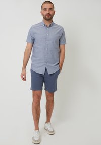 Camisa de botones de manga corta color azul claro con cuello, combinada con pantalones cortos navy y zapatillas ligeras. La tela parece suave y liviana.