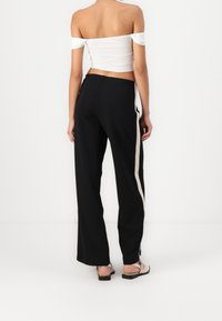 Kobieta ubrana w czarne szerokie spodnie z białymi bocznymi paskami oraz białą bluzkę typu crop off-shoulder, stojąca na jednolitym białym tle.