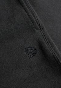 Sort bomuldsstof med en subtil struktur, med et præget logodesign i nederste venstre hjørne. Ingen lynlåse eller ekstra hardware synlige.