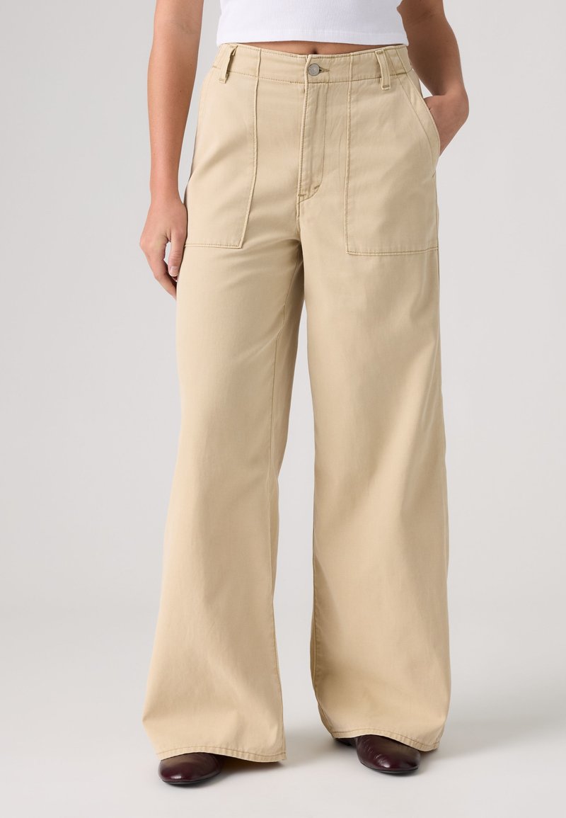 Femme portant un pantalon beige à jambes larges avec poches plaquées à l'avant, un top blanc court et des chaussures foncées, debout devant un fond uni.