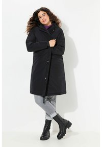 Zwarte waterdichte jas met een capuchon, voorkeursrits, zijzakken en een knielange ontwerp, gedragen over grijze broek en zwarte laarzen.