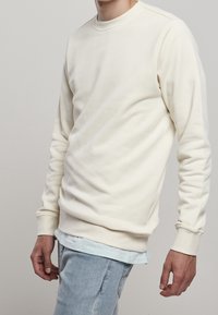 Sudadera color crema con cuello redondo, mangas largas y puños de canalé, usada sobre una camisa con capas de color azul claro, de tela texturizada.