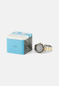 Montre Fossil avec un cadran noir, un bracelet en acier inoxydable doré et argenté, et une lunette noire. Livrée avec une boîte de rangement bleue et à motifs.
