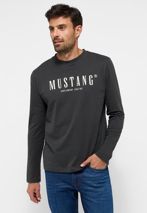 Gray long-sleeve t-shirt with "MUSTANG" logo in cream. Cotton blend fabric, crew neck design, worn with blue jeans.