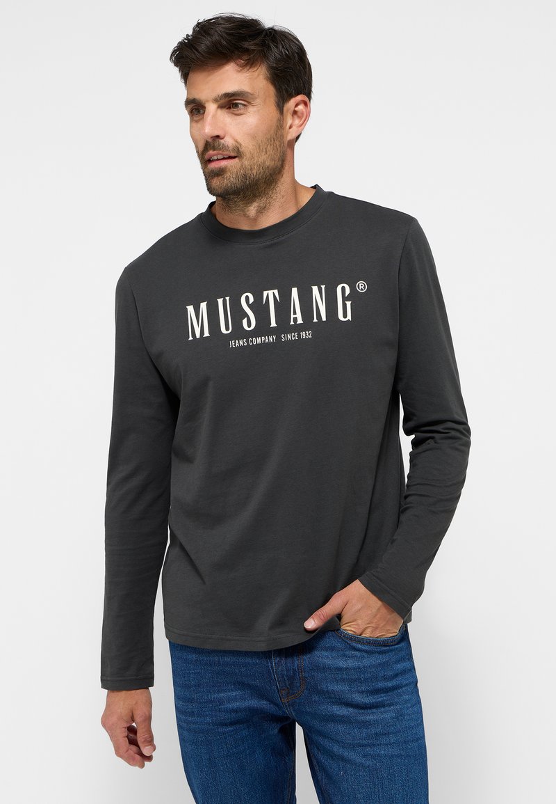 Pilkas ilgomis rankovėmis marškinėliai su "MUSTANG" logotipu kremine spalva. Medvilnės mišinys, apvalios kaklo iškirpės dizainas, derinamas su mėlynomis džinsinėmis kelnėmis.