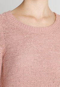 Nahaufnahme eines hellrosa gestrickten Pullovers mit runder Ausschnitt, der von einer Person getragen wird, wobei der Hals- und obere Brustbereich zu sehen ist.