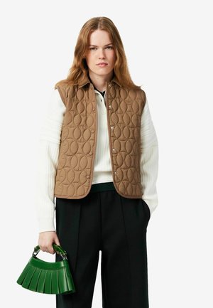 Jonge vrouw met lang haar die een bruin doorgestikt vest, een witte trui, een zwarte broek draagt en een kleine groene handtas vasthoudt.