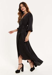 Black wrap dress with long, puffed sleeves, silky texture, and a tied waist. Features a high-low hem and is paired with chunky black heels.