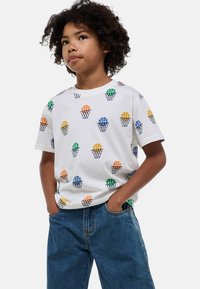 T-shirt blanc à manches courtes avec des motifs colorés de basketball et de panier en vert, orange, bleu et jaune, associé à un jean en denim.