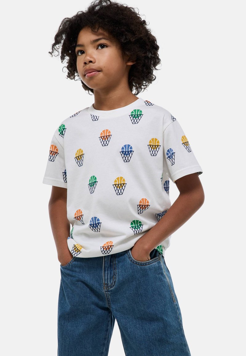 T-shirt blanc à manches courtes avec des motifs colorés de basketball et de panier en vert, orange, bleu et jaune, associé à un jean en denim.