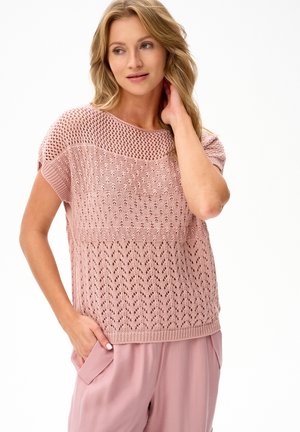 Kobieta w luźnym, różowym krótkim sweterku z różnymi wzorami koronkowymi i pasujących różowych spodniach, pozująca z jedną ręką w kieszeni.