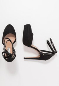 Sorte høyhælte sko med plattform, laget av semsket skinn, tynne ankelremmer og spisse tær. Hælen er stiletto-stil med en blank finish.