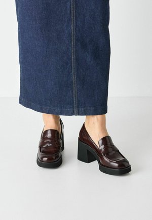 Mocassini in pelle bordeaux scuro con tacco alto nero, caratterizzati da punta arrotondata e dettagli di cucitura sottili. Abbinati a una gonna di jeans.