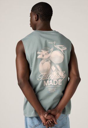 Hombre visto desde atrás, con camiseta sin mangas verde con gráfico de durazno y texto "Tastefully Made", manos entrelazadas detrás, vaqueros azul claro.