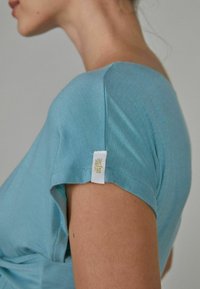 Korte mouwen, lichtblauwe top van zachte stof. Bevat een klein label op de mouw met gouden tekst. Zachte textuur, fitted ontwerp.