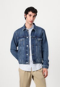 En mørkeblå denimjakke med klassisk snitt, med knappelukking foran, to brystlommer, og kontraststing, båret over en stripete skjorte.