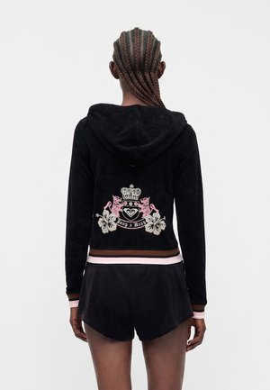 Personne aux cheveux tressés portant une veste à capuche noire avec un blason floral rose et blanc et un short noir, vue de dos.