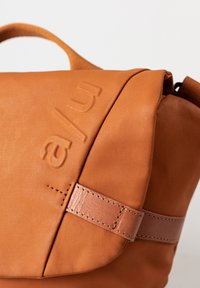 Borsa messaggera color sabbia con superficie testurizzata, logo impresso e dettagli in pelle. Presenta una chiusura a patta e cuciture decorative.