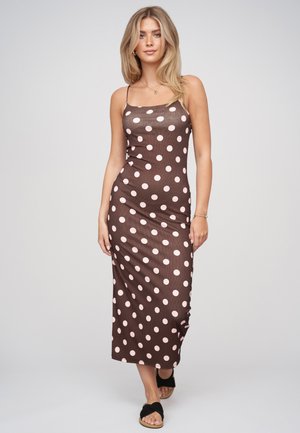 Donna con capelli biondi che indossa un vestito marrone senza maniche con pois bianchi e sandali neri con dita aperte, in piedi davanti a uno sfondo chiaro uniforme.