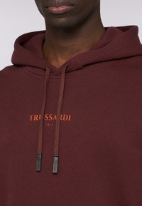 Bordowy hoodie z teksturowanej tkaniny, z regulowanym sznurkiem zakończonym metalowymi końcówkami oraz pomarańczowym logo na piersi.