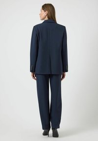 Tmavě námořnický oblek na míru se slabě oversized sakem, rovnými kalhotami, hladkou texturou a decentními knoflíkovými detaily na rukávech.