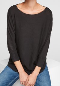 Czarny, luźny sweter z szerokim dekoltem i rękawami do trzech czwartej, wykonany z miękkiego, lekkiego materiału. Noszony z niebieskimi dżinsami.