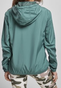 Lekka, niebieskozielona kurtka z kapturem, elastycznymi mankietami i wciąganym pasem. Gładka tekstura, odpowiednia do użytku na zewnątrz. Częściowo zakryte legginsy w kamuflażu.