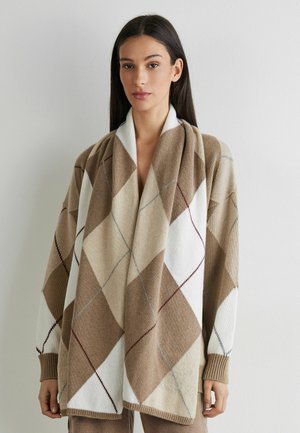 Femme portant un cardigan en maille beige et blanc oversize à motif losanges, manches longues, debout contre un mur clair uni.