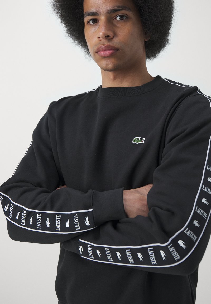 Schwarzer Pullover mit Rundhalsausschnitt, ausgestattet mit weiß gestreiften Ärmeln. Kleines grünes Lacoste-Logo auf der Brust, mit wiederholtem Logo-Muster auf den Ärmeln.