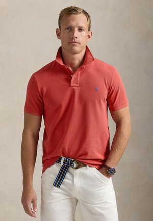 Svetlolas moški, oblečen v polo majico s salmonovo rdečo barvo in ovratnikom dvignjenim navzgor, bele hlače, pas s črtami in ročno uro, stoji pred enobarvnim ozadjem.