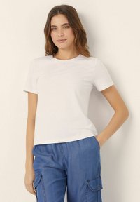 Tricou alb cu mâneci scurte și decolteu rotund, realizat din bumbac neted, asortat cu pantaloni cargo albaștri cu buzunare laterale.