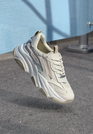 Baskets blanches épaisses avec détails en mesh et cuir, logo "Steve Madden" sur le côté, flottant au-dessus d'une surface pavée grise.