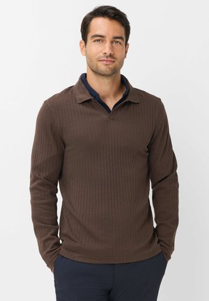 Uomo con capelli scuri che indossa una polo marrone a coste a maniche lunghe e pantaloni scuri, in piedi con le mani nelle tasche su sfondo bianco.