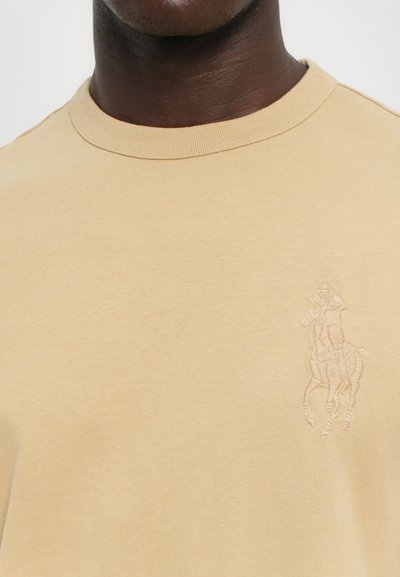 Beige bomull t-shirt med rund halsringning, med en subtil broderad hästsportdesign på det vänstra bröstområdet. Slät textur.