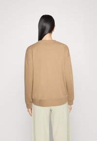 Brązowy oversized sweter z okrągłym dekoltem i ściągaczami, charakteryzujący się miękką fakturą i minimalistycznym designem, noszony z jasnymi spodniami.