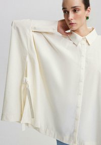 Chemise en coton blanche à coupe décontractée, avec un col, des boutons sur le devant et des manches dramatiques en forme de cape avec des liens.