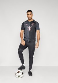 Svart träningsskjorta med rosa logga och detaljer, tillsammans med svarta byxor med vita sidostriped. Svart fotboll vid fötterna.
