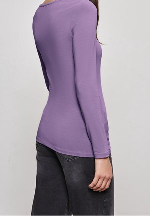 Donna che indossa un top aderente a maniche lunghe di lavanda e jeans grigio scuro, in piedi contro uno sfondo grigio chiaro uniforme.