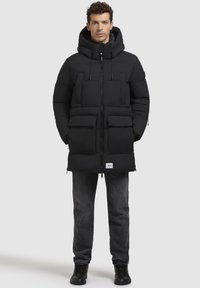 Doudoune noire avec capuche, dotée de deux poches avant, de zips et de cordons de serrage réglables ; associée à un jean foncé et des chaussures noires.