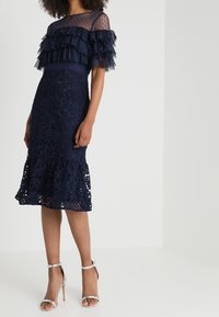 Robe bleu marine présentant un haut transparent superposé à volants avec des pois, une jupe en dentelle élaborée et des escarpins à brides argentées à la cheville.