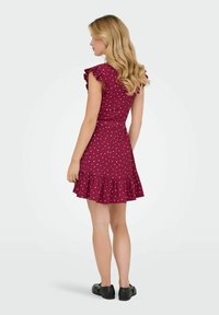 Burgundy prickigt klänning med korta volangärmar, figurnära midja och utsvängd nederkant. Bärs med svarta skor, som visar en texturerad tyg.