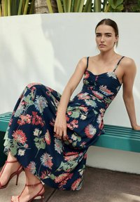 Marineblaues Maxikleid mit Blumenmustern, dünnen Trägern und gerafftem Oberteil. Getragen mit roten Riemchenheels, sitzend auf einer türkisen Bank.