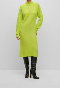 Robe en maille à manches longues de couleur vert citron vif, dotée d'un col montant et d'une coupe décontractée, associée à des bottes en cuir noir au-dessus du genou.